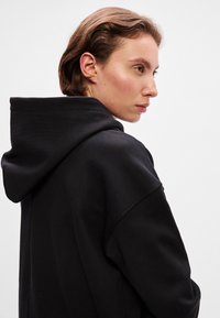 Musta huppari rentoa istuvuutta, pehmeää kangasta ja näkyviä saumoja. Sisältää suuren, pyöreän huppukappaleen ja laskeutuvat olkaimet.