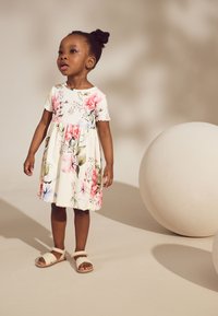 Rochie florală cu mâneci scurte, bază albă, flori roz și albastre. Tiv texturat. Sandale căprui cu bretele. Fundal neted, neutru.