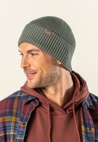 Olive green knitted beanie with ribbed texture, featuring a small brown tag. Worn with a brown hoodie and plaid flannel shirt.