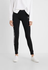 Svarta leggings med en åtsittande silhuett, med en vit tryckt logotyp på vänster ben. Tillverkade av stretchigt material, tillsammans med vita sneakers.