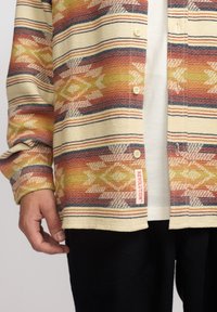 Man in een multicolor gestreepte blouse over een wit T-shirt met zwarte broek, hand ontspannen aan de zijde, label "REVOLUTION" zichtbaar.