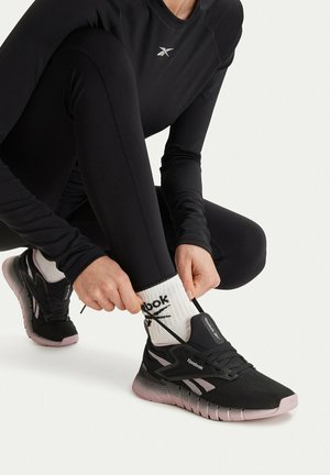Persona con ropa deportiva negra atándose unas zapatillas Reebok negras y rosas con calcetines blancos de Reebok sobre fondo blanco.