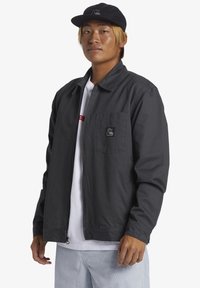 Black zip-up jacket with a collar, front pocket, and logo patch. Worn over a white shirt and paired with light-blue pants and black cap.