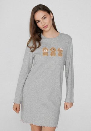 Joven mujer vistiendo un vestido gris de manga larga con tres diseños de perrito de peluche en el pecho, sonriendo suavemente con el cabello suelto.