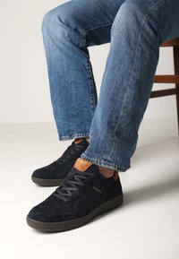 Sneakers in suede nero con dettagli testurizzati, dettagli del tallone marroni e suole in gomma scura, abbinati a jeans in denim blu.