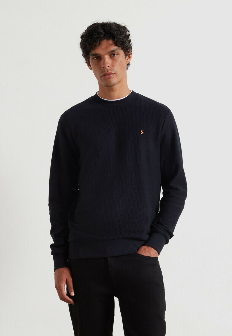 Marineblauw sweatshirt met een textuurpatroon, ronde halslijn en geborduurd logo in oranje. Aansluitende manchetten en tailleband, gecombineerd met zwarte broeken.