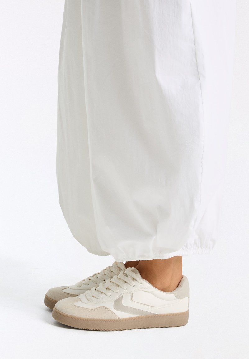 Baskets décontractées blanches et beiges portées avec un pantalon blanc ample, vues de profil sur un fond blanc.