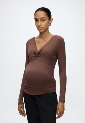 Gravid kvinde iført en tætsiddende brun langærmet top med en opsamlet detalje ved brystet og sorte bukser, stående mod en ensartet baggrund.