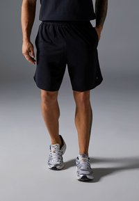 Svarta sportshorts med elastisk midja, sidofickor och avslappnad passform, kombinerade med grå och vita löparskor.