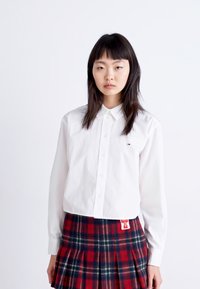 Camisa blanca de cuello con botones, combinada con una falda de cuadros rojos con pliegues que presenta acentos en verde y azul y una etiqueta.