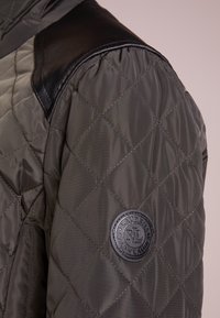 Lapptäckt jacka i olivgrön med svarta läderaxelaccenter och en cirkulär logotyp som har "Ralph Lauren" på vänster ärm.