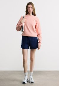 Νέα γυναίκα που φοράει ροζ μακρυμάνικο φούτερ, σκούρα μπλε σορτς, λευκές κάλτσες και λευκά αθλητικά παπούτσια, κρατώντας μια ασημένια τσάντα ώμου στον ώμο της.