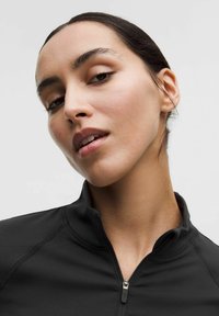 Must mustriibuga must sportlik ülemine osa kõrge kaelusega, sujuva tekstuuriga, nähtava õmblusega ja metallist lukuga. Mudeli nägu osaliselt nähtav.