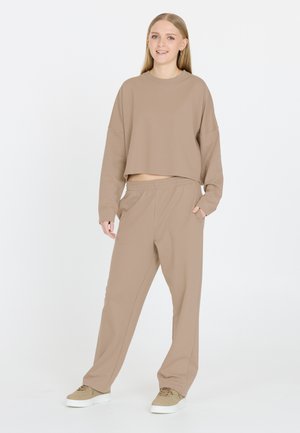 Nuori nainen seisoo, pukeutuneena beigeen löysään svetariin ja vastaaviin housuihin, beigeihin lenkkareihin, toinen käsi taskussa valkoista taustaa vasten.