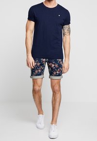 Marinblå crew-neck T-shirt med en liten bröstlogotyp, kombinerad med shorts i blommönster med uppvikta muddar. Vita sneakers fulländar outfiten.