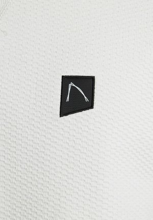 Tessuto bianco testurizzato con una piccola toppa nera caratterizzata da un design geometrico bianco. La toppa ha un bordo irregolare, creando un contrasto visivo.