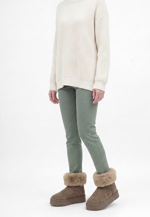 Personne portant un pull en maille crème, un pantalon slim vert et des bottines marron doublées de fourrure, debout sur un fond blanc.