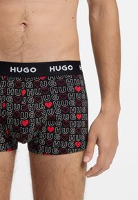 Sorte boksertrusser med flerfarvet "HUG"-tekst og røde hjerter som mønster. Har en kontrasterende sort elastik med "HUGO"-logo.