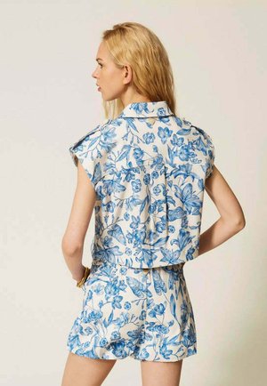 Donna bionda vista da dietro che indossa una canottiera bianca e pantaloncini con motivo floreale blu, in piedi davanti a uno sfondo uniforme.