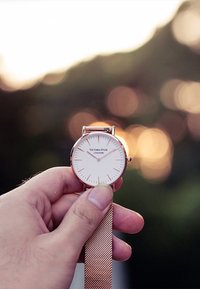 Orologio rotondo con quadrante bianco, cassa in oro rosa e cinturino a rete. Presenta indicatori delle ore sottili e un design minimalista. Tenuto in mano davanti a uno sfondo bokeh.