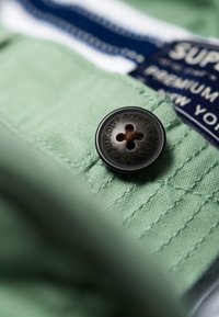 Groene katoenen stof met een donkerbruine knoop met vier gaten en gegraveerde tekst. Bevat een blauw label met witte letters.