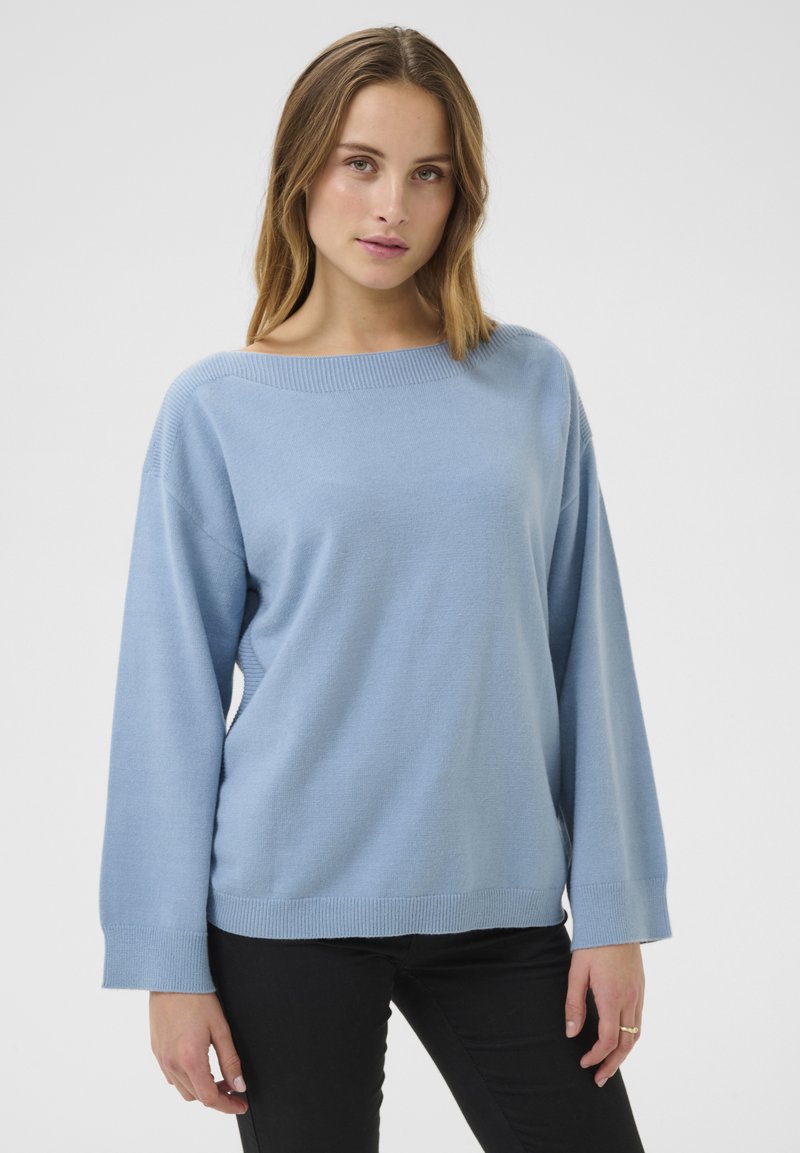 Maglione azzurro chiaro a maglia con scollo ampio e maniche lunghe, caratterizzato da una morbida texture e polsini a coste. Indossato con pantaloni neri.