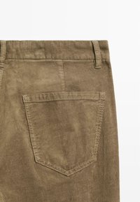 Bruine corduroy stof met verticale ribbels. Bevat een achterzak met stiksels en een standaard tailleband. Gekartelde structuur.