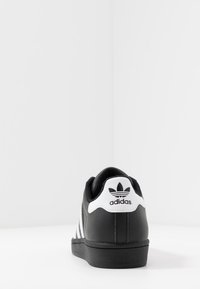 Černá kožená teniska s bílými akcenty, design se třemi pruhy, polstrovaný límec a černá gumová podrážka. Logo Adidas je vzadu.