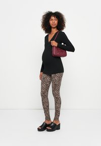 Femme enceinte portant un haut noir, un legging à imprimé léopard, des sandales à talons noires, tenant un sac à main bordeaux sur son épaule, se tenant contre un fond blanc.