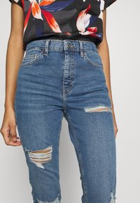 Vaqueros de denim azul con cintura alta, que presentan detalles desgastados y roturas en las rodillas. Bolsillos estándar y cierre clásico de botón.