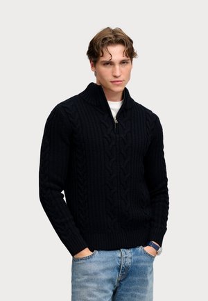 Jeune homme aux cheveux ondulés portant un pull zippé noir en maille torsadée sur une chemise blanche et un jean bleu, les mains dans les poches, sur un fond uni.