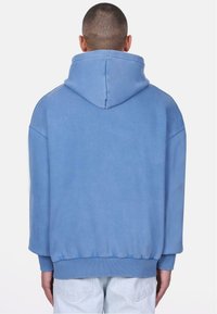 Blå sweatshirt med hætte, blød tekstur, afslappet pasform, ribbede manchetter og kant, samt en glat overflade. Bagfra viser den et minimalistisk design.