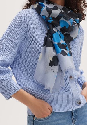 Person trägt eine hellblaue Strickjacke mit großen Knöpfen und einen schwarz-blau-grauen gemusterten Schal, Hand in der Tasche, beschnittener Blick.