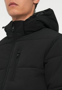 Sort foret nedjakke med lynlås på forsiden, høj krave og justerbar hætte. Glat tekstur med minimalistisk design og diskret lynlås lomme.