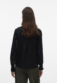 Zwarte blouse met lange mouwen en gestructureerde stof, voorzien van subtiele patronen en geplooide manchetten. Gecombineerd met olijfgroene broek, gezien van de achterkant.