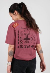 T-shirt en coton bordeaux avec un design graphique noir comprenant des formes et des motifs abstraits à l'arrière ; comprend une étiquette rouge sur l'épaule.