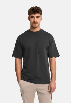 Homme aux cheveux bruns bouclés portant un t-shirt à manches courtes gris foncé avec une poche poitrine et un pantalon cargo beige, debout les mains dans les poches.