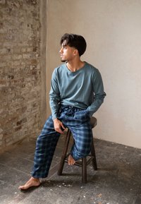 Tričko s dlouhým rukávem v barvě šalvěje spárované s tmavě modrými károvanými pyžamovými kalhotami s elastickým pasem, zobrazené vsedě na dřevěné stoličce.