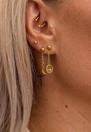 Ohr mit mehreren goldenen Piercings, darunter ein halbmondförmiger Anhänger und ein gedrehter Kreis, die an Ketten hängen, und ein kleines Kreuz-Tattoo hinter dem Ohr.