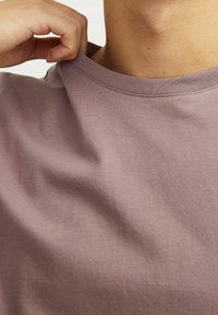Mauve pamučna majica s okruglim izrezom i kratkim rukavima. Tkanina ima glatku teksturu i sadrži suptilne nabore blizu ovratnika.