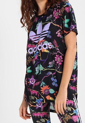 Personne portant un t-shirt noir Adidas et un legging avec des motifs floraux et papillons colorés, avec un grand logo Adidas violet pastel.