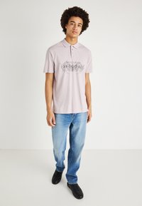 Ροζ πόλο μπλούζα με γιακά και μαύρο γραφικό σχέδιο μπροστά, συνδυασμένη με ξεθωριασμένα μπλε τζιν και μαύρα αθλητικά παπούτσια.