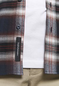 Koszula flanelowa z granatowym, czerwonym i białym wzorem w kratę, z czarnymi guzikami i metką "Peak Performance", noszona na białym T-shircie.