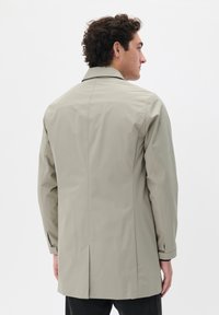 Impermeable de color beige claro con un diseño recto, que presenta un cuello clásico, mangas largas y una abertura en la parte trasera, fabricado con un material suave y a prueba de agua.