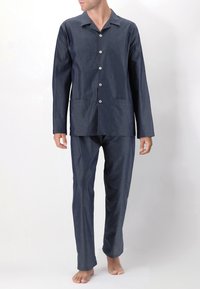 Uomo in piedi a piedi nudi, che indossa un pigiama blu navy con maniche lunghe, bottoni e piccoli motivi a pois bianchi, abbinato a pantaloni.