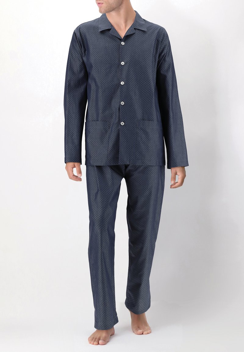 Uomo in piedi a piedi nudi, che indossa un pigiama blu navy con maniche lunghe, bottoni e piccoli motivi a pois bianchi, abbinato a pantaloni.