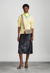 Κίτρινος πλεκτός σκούφος, ναυτικό μίντι φούστα με παγιέτες, μπεζ τσάντα ώμου με λογότυπο και καφέ ψηλοτάκουνα σανδάλια. Πολυάριθμα κολιέ με χρώματα.