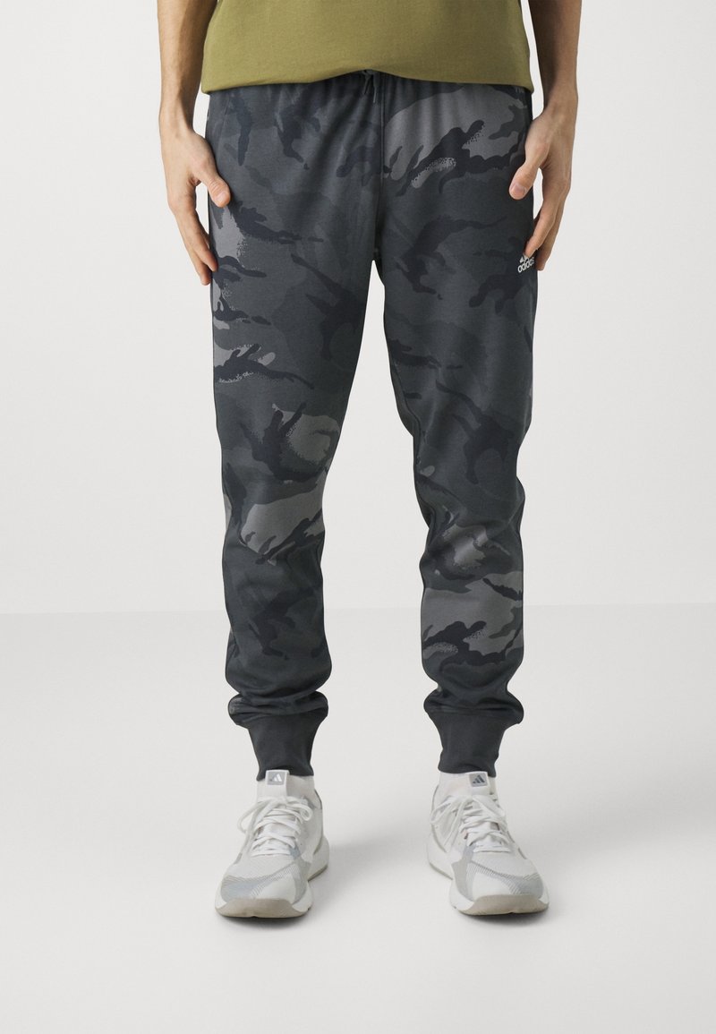 Kamouflagebyxor i mörkgrå nyanser med dragsko, ribbade muddar och sidofickor, kombinerat med ljusgrå sportskor.