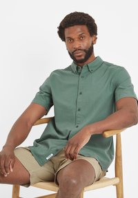 Short-sleeved, button-up shirt in dark green fabric with a smooth texture. Features a collar and side label. Worn with light beige shorts.