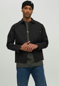 Sort jakke med knaplukning, brystlomme og tekstureret stof, lagd over en grøn skjorte og blå jeans.
